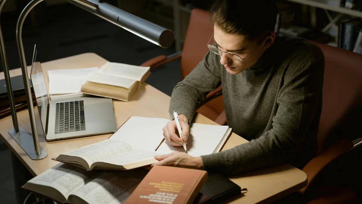 A student at a desk taking notes from books under warm lamp light.