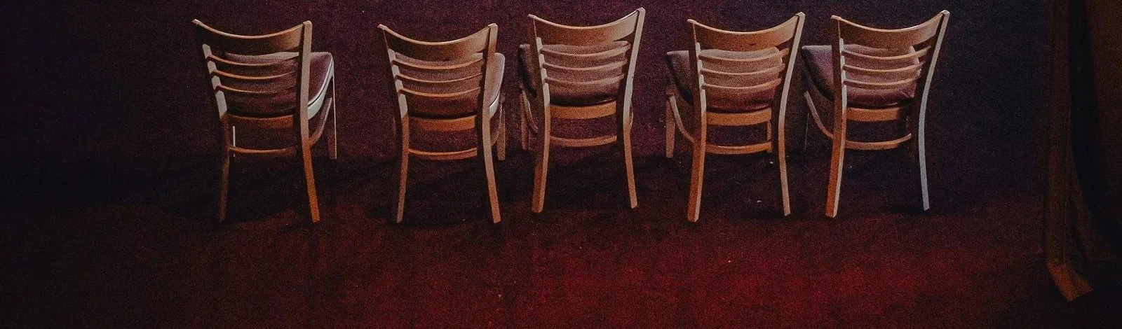 A row of empty wooden chairs in a deep red theatrical room with a small lit stage in the background.