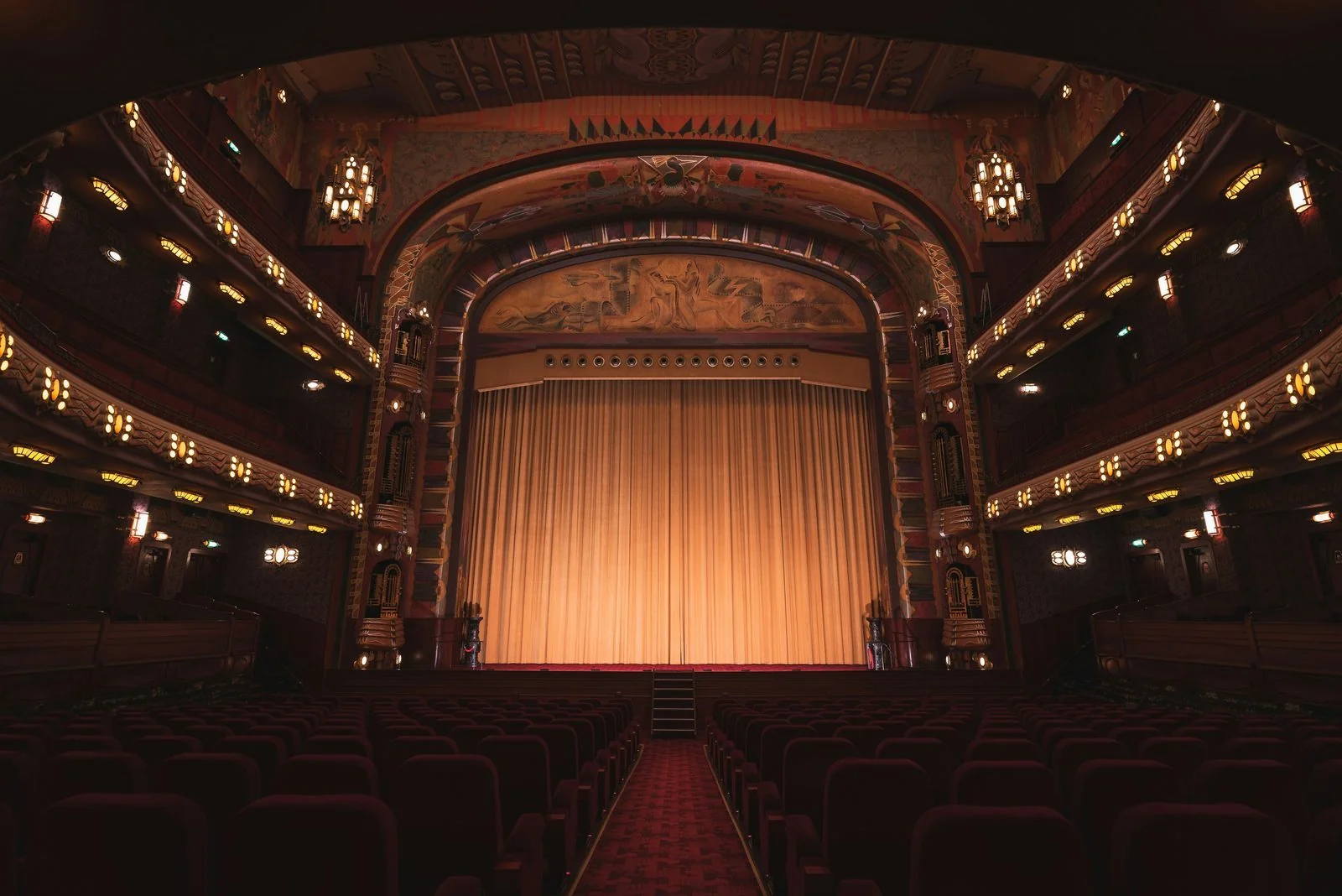 A historic theatre interior with a closed gold curtain and ornate ceiling, viewed from the empty seats.